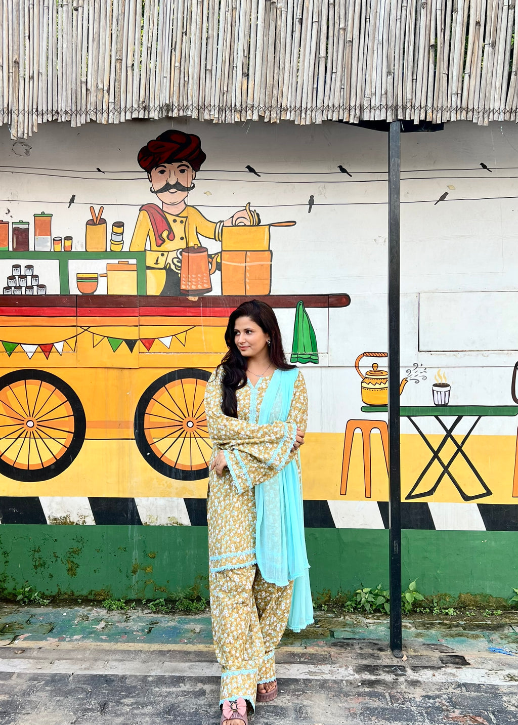 A woman modeling a traditional South Asian outfit consisting of a beige and yellow floral print kurta and trousers with a light blue dupatta, standing in front of a colorful painted wall depicting a street food vendor.